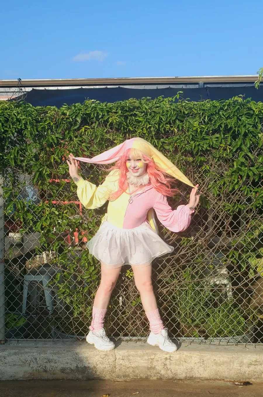 Clown performer Sleepie Circus in a pink and yellow clown outfit. She's standing in front of a brick wall and posing for the camera.