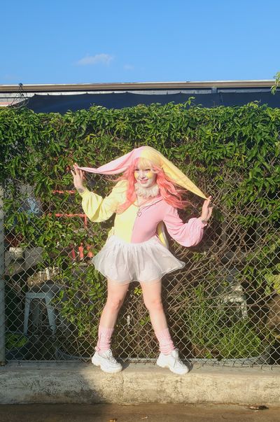Clown performer Sleepie Circus in a pink and yellow clown outfit. She's standing in front of a brick wall and posing for the camera.
